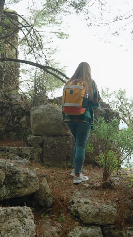 Vertical video. A woman with a backpack walks through the forest and explores ancient ruins of houses that were destroyed thousands of years ago.