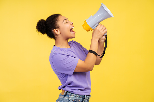 Discount news theme. Image of a beautiful young African American female in a t-shirt, screaming into a loudspeaker, on a vivid yellow background