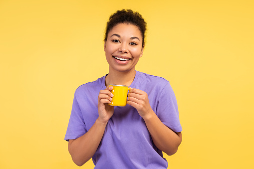 Human Emotions Concept. Photo of a delightful smiling young African American woman in a t-shirt, holding a mug of coffee, isolated on a yellow background. Morning energy theme