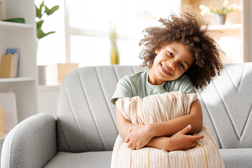 Immersed in a relaxed home ambiance, an African American girl perches on a sofa with a pillow in arms, her beaming smile portraying a scene of comfort and simplicity