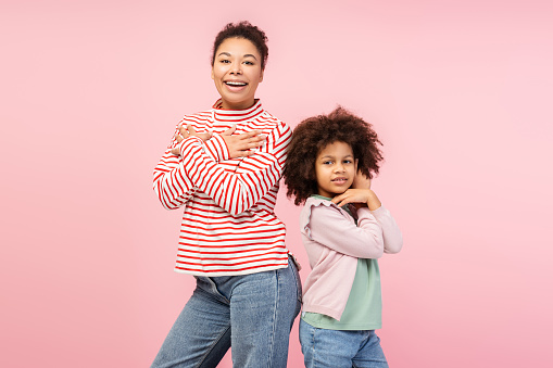 Playful family photo idea. Shot of a radiant African American mom and her daughter, posing and laughing together, captured isolated on a pastel pink background. Casual, fun-filled ambiance