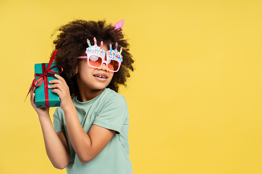 Birthday happiness theme. Photograph of radiant little African American girl excited about her gift. She dons playful party glasses and holding present near her ear isolated on vivid yellow background