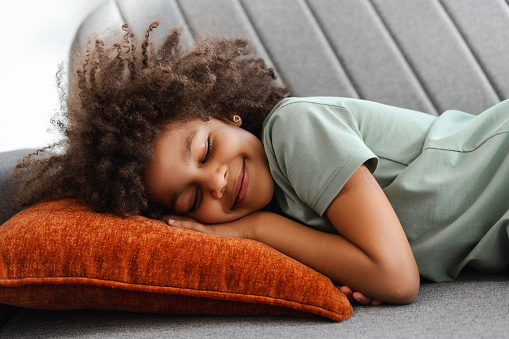 Picture of a delightful African American young girl reclining on a sofa pillow, her bright smile showcasing the warmth and comfort of a casual home atmosphere. Cozy home interior concept