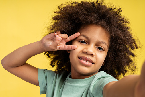 Joyful emotions captured. Image of little African American girl with curly hair, taking selfie. She gazes into camera, displaying victory sign with her fingers, isolated on yellow background