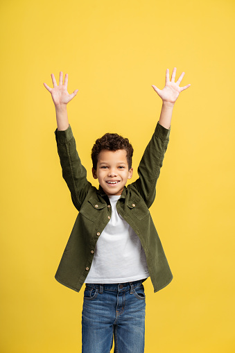 Capture of an animated young african american boy in fashionable casuals, springing with hands up, facing the camera, against a lively yellow background. Children's fashion concept on vertical photo