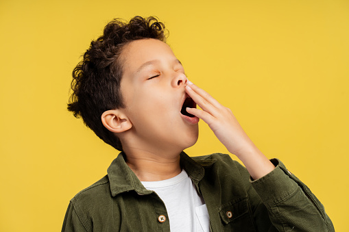 Youth sentiment concept. Close view of a little fatigued african american boy dressed in contemporary fashion, yawning and placing his hand over his mouth, isolated on a bright yellow background