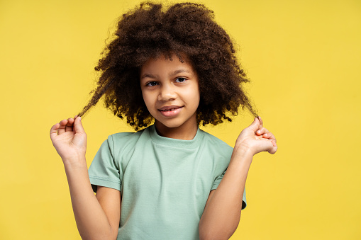 Bright-eyed African American girl with curly strands, dressed in a light mint t-shirt, lifts her hands, gently pulling at her locs with a joyful expression, isolated on a lemon-yellow background