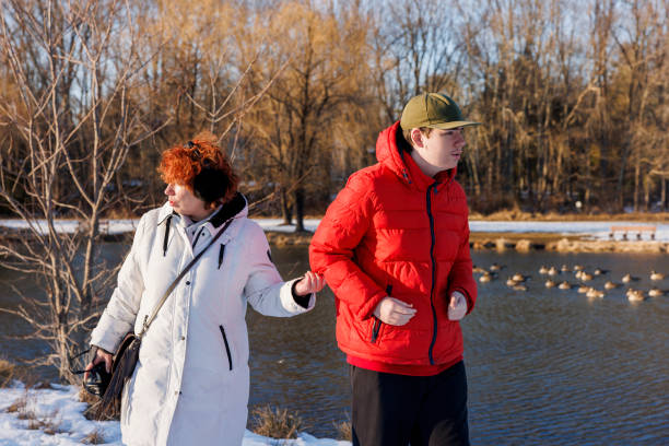 Sociable Autistic young man in warm clothing, strolls with mom through winter park by the lake with ducks