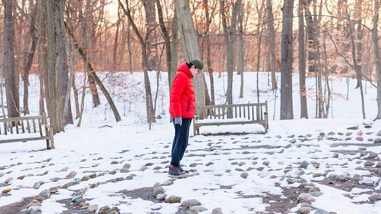 Lonely young Autistic man in a jacket and cap carefully makes his way through the stone spiral maze in snowy park