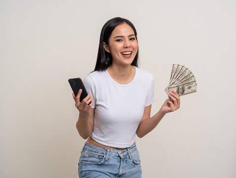 Cheerful Rich young Asian business woman showing lot of money and smartphone. Attractive female with dollar banknotes. Happy asian woman  counting money us dollar on isolated white background