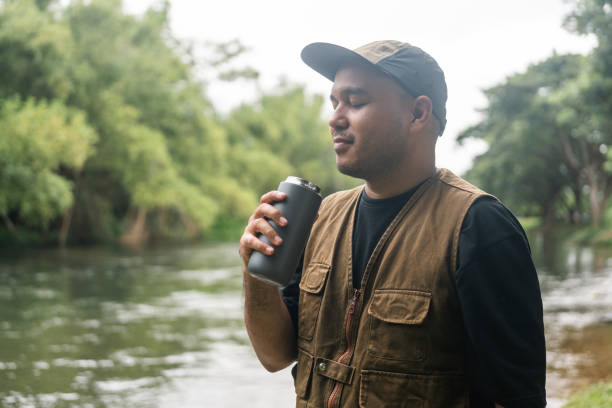 Young asian man traveller relaxing drinking coffee in nature forest and river background. Happy male living in middle of a lot of tree. Breathing the fresh air on her vacation weekend holiday trip.