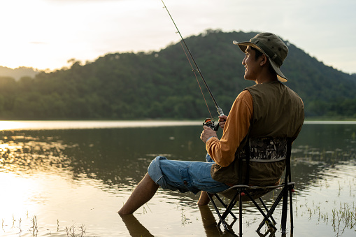 Enjoy moment of Handsome man fishing as a leisure activity during his vacation at the lake on sunset. Silhouette at sunset moment of man fishing rotation with reel.