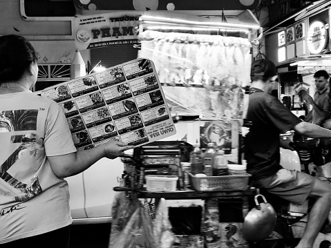 Local lady with restaurant menu and ambulant seller in Bui Vien Street, the main nightlife street of Pham Ngu Lao, the so called ‘backpackers area’ of Ho Chi Minh City, Vietnam