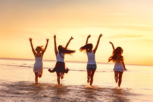 A group of happy young girls are running along the calm sunset beach of a lake in shallow water