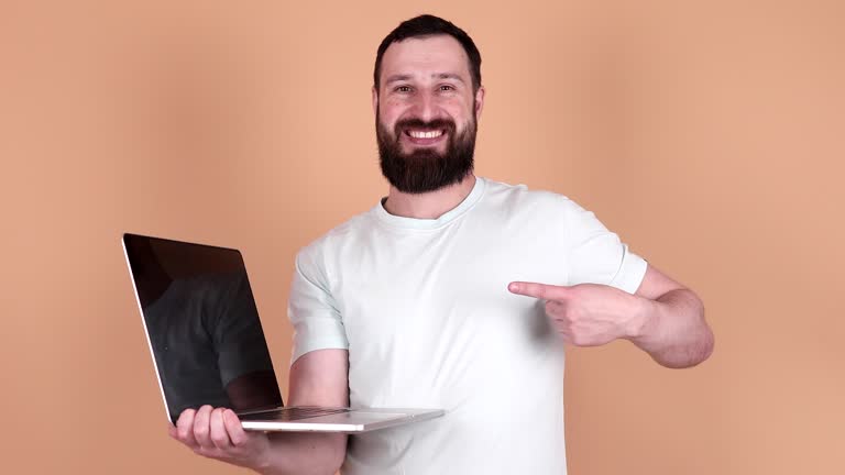 Young happy bearded caucasian IT man he wear shirt hold in hands typing use work point index finger on laptop pc computer isolated on plain beige background studio portrait.