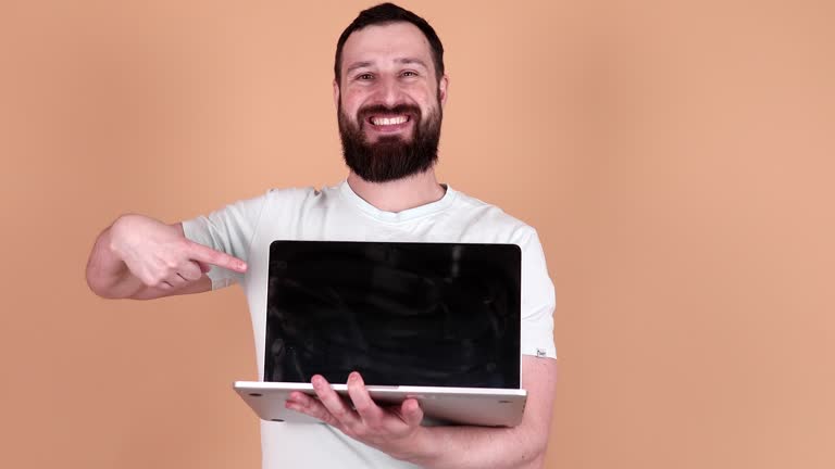 Young happy bearded caucasian IT man he wear shirt hold in hands typing use work point index finger on laptop pc computer isolated on plain beige background studio portrait.
