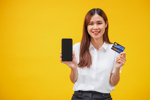 Smiling young Asian woman  pointing with smartphone and  credit card Shopping online standing on yellow background.