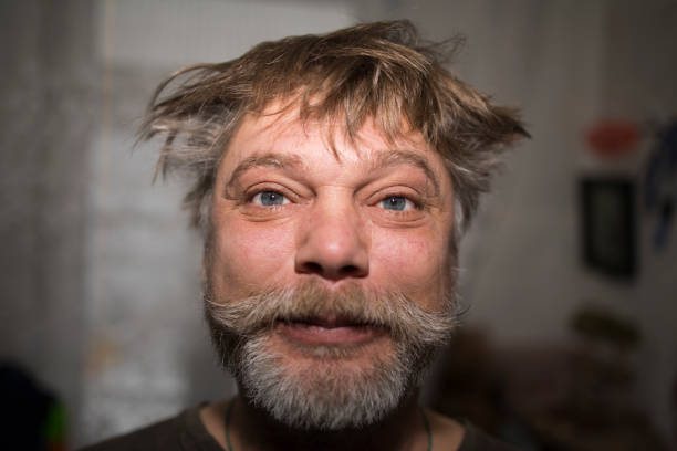 A man with a full beard and disheveled hair is gazing up at the camera, revealing his forehead, nose, slight smile, jawline, neck, ears, and facial hair including a moustache