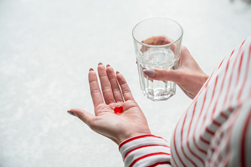 Red pill on a female palm of hand and glass of water in their hand. A woman taking a medicines at home.