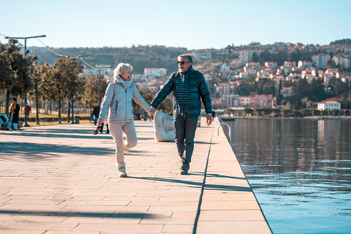 A senior Caucasian male and female couple enjoy a leisurely walk along a sunlit seaside promenade with a view of a coastal town in the background.