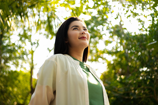 A Adult Asian woman with a serene expression embraces the warmth of the sunlight, surrounded by the vibrant greenery of a peaceful garden.