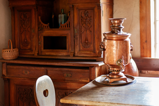 Interior of an ancient guest room. In the room there is a beautiful carved wood cabinet for dishes, a sunlit table and a copper Russian kettle for tea on it.