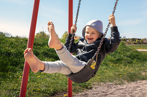 A happy toothless boy raises his bare feet and swings hard on a chain swing in the playground.