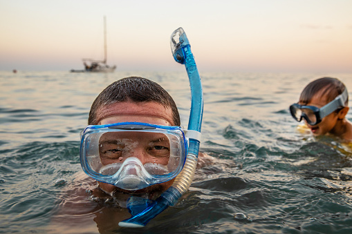 Portrait of a young cheerful Caucasian man in a mask and snorkel, who swims in the warm evening sea after sunset from his sailboat with his little 3 year old son. Father and son have fun and dive.