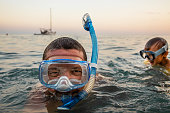 Portrait of a young man swimming in the sea from his yacht at sunset snorkeling with his little son who is also wearing a swimming mask.