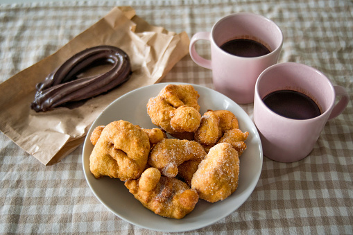 Bowl of Buñuelos Fritters with Sprinkled Sugar next to Big Chocolate Churro and Two Pink Cups of Liquid Hot Chocolate. Typical Valencian Breakfast in Fallas