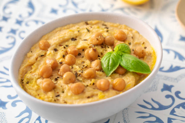 A bowl of hummus with chickpeas and basil is displayed on a table, showcasing a mix of Food ingredients. Tableware adds to the presentation of this delicious Cuisine
