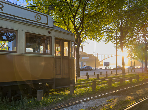 PORTO, PORTUGAL - CIRCA APRIL 2023: Wooden historical vintage yellow tram 287 moving on Porto street, symbol of city.Old tram passing by in Massarelos with the Arrabida bridge in the background during sunset. Indispensable transport for locals and interesting attraction for tourists. Portugal