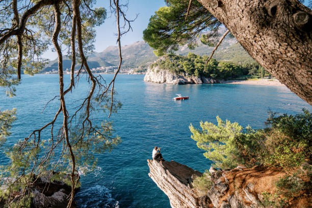 View through the green branches on a man sitting on a stone ledge and looking at a boat passing by the shore. Back view