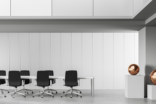 Semi-empty office interior with a large blank white wall with copy space on the gray concrete floor. Work desks with chairs and computer equipment, potted plants on top of the desks, black pendant lights, and windows with a garden view in the background. 3D rendered image.