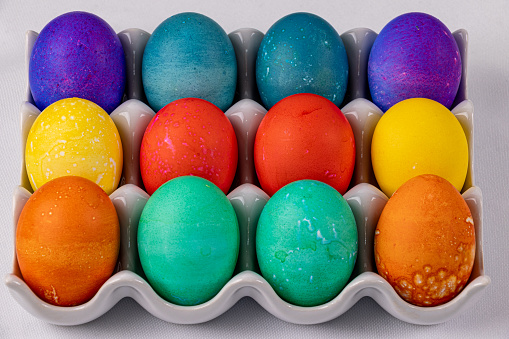 Close up of twelve brightly colored eggs of yellow purple green red orange and yellow in a white ceramic dish