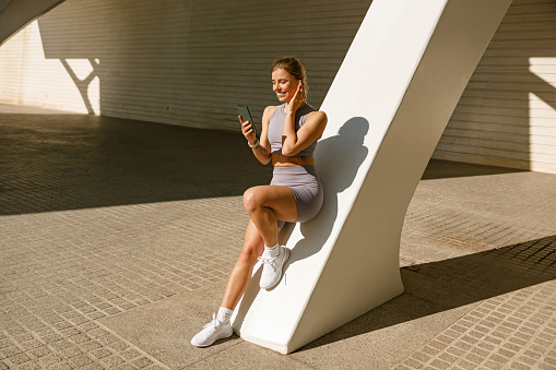 Fit woman in sportswear standing outdoors after training and use mobile phone