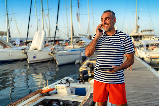 Mature man standing on the dock dressed in a sailor's t-shirt and speaking a phone