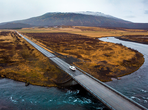 Aerial drone captures a scenic view of a car driving over a bridge in Iceland, surrounded by breathtaking mountainous landscape and serene waters below