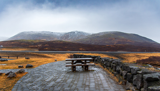 A serene picnic spot awaits amidst the stunning natural beauty near Thingvellir national park in Iceland, with a wooden bench on a stone walkway overlooking snow-capped mountains and a tranquil lake in the background