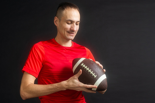 Young man dressed in casual street clothes holds a football, ready to throw a pass, isolated on black background. High quality photo