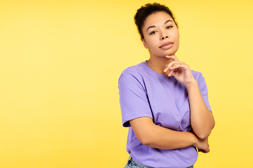 Concept of allure. Captivating young African American female in a casual outfit, gently caressing her jawline with her finger, looking intently at the camera, isolated on a yellow background