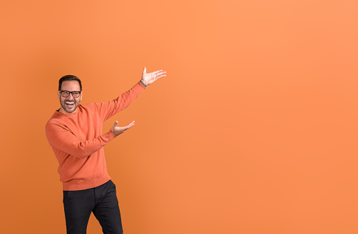 Ecstatic businessman in eyeglasses with arms raised advertising on copy space over orange background