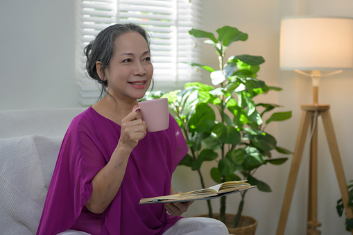 Joyful and relaxing Asian senior woman enjoy her delicious cup of coffee while relaxing on the sofa at cozy home.