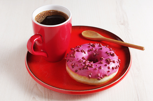Pink donut with sugar balls and a cup of coffee on a red dish on a wooden table