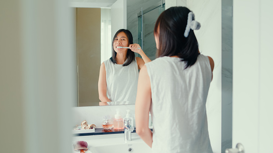 Young Asian woman having fun use toothbrush brush teeth in front of mirror in lavatory at home late morning. Routine lifestyle in house concept.