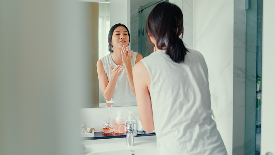 Young Asian woman having fun apply facial cream look in mirror in lavatory at home late morning. Routine lifestyle in house concept.