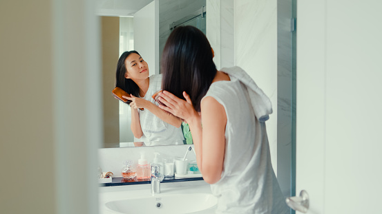 Young Asian woman having fun combing her beautiful hair in front of mirror in lavatory at home late morning. Routine lifestyle in house concept.