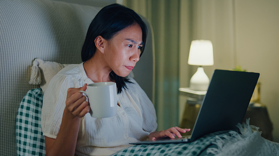 Young Asian girl using laptops and drinking milk sitting on the bed in home at night. Insomnia, Cybersickness, Nomophobia, sleep disorder concept.