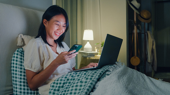 Young Asian girl using laptops and phone sitting on the bed in home at night. Insomnia, Cybersickness, Nomophobia, sleep disorder concept.