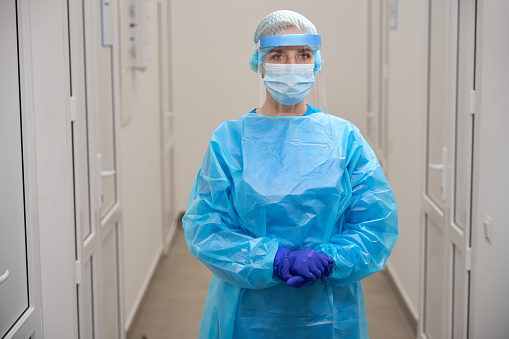 Female infectious disease specialist stands in a bright corridor in protective equipment, she uses a protective shield